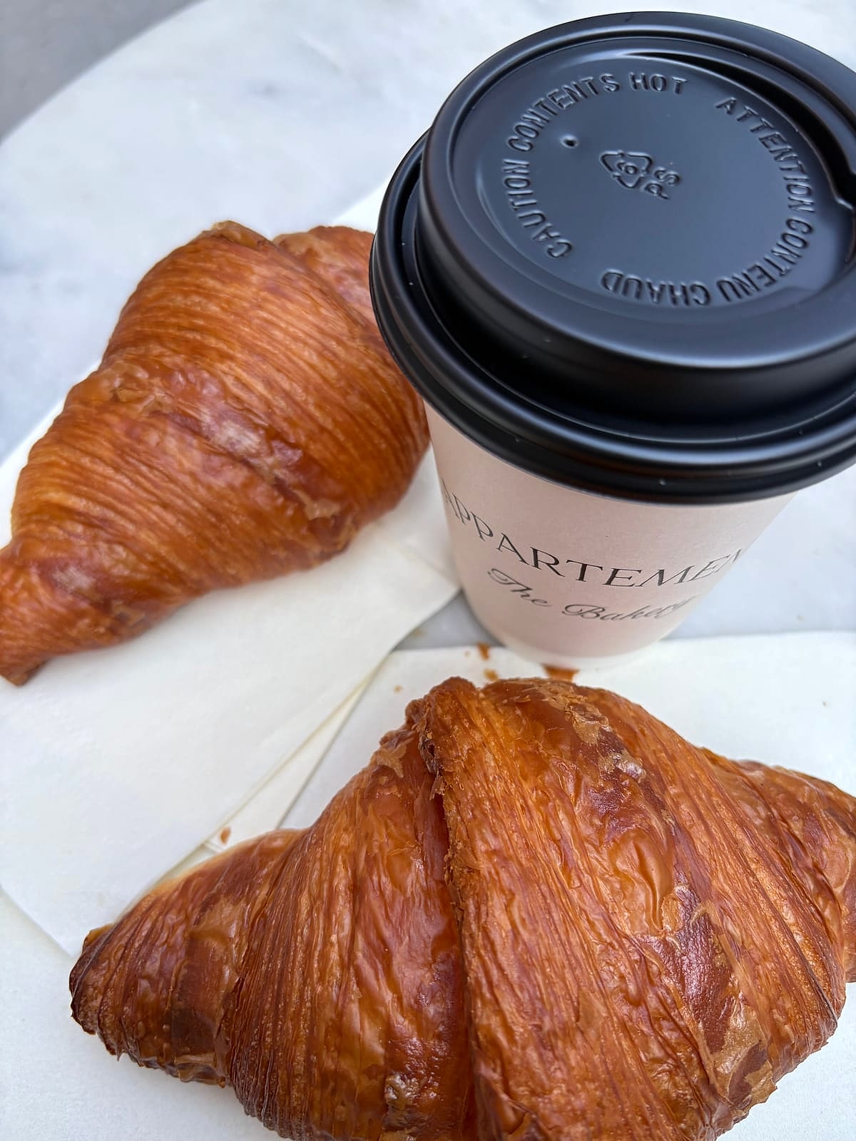 Two flaky croissants next to a L'Appartement 4F coffee cup